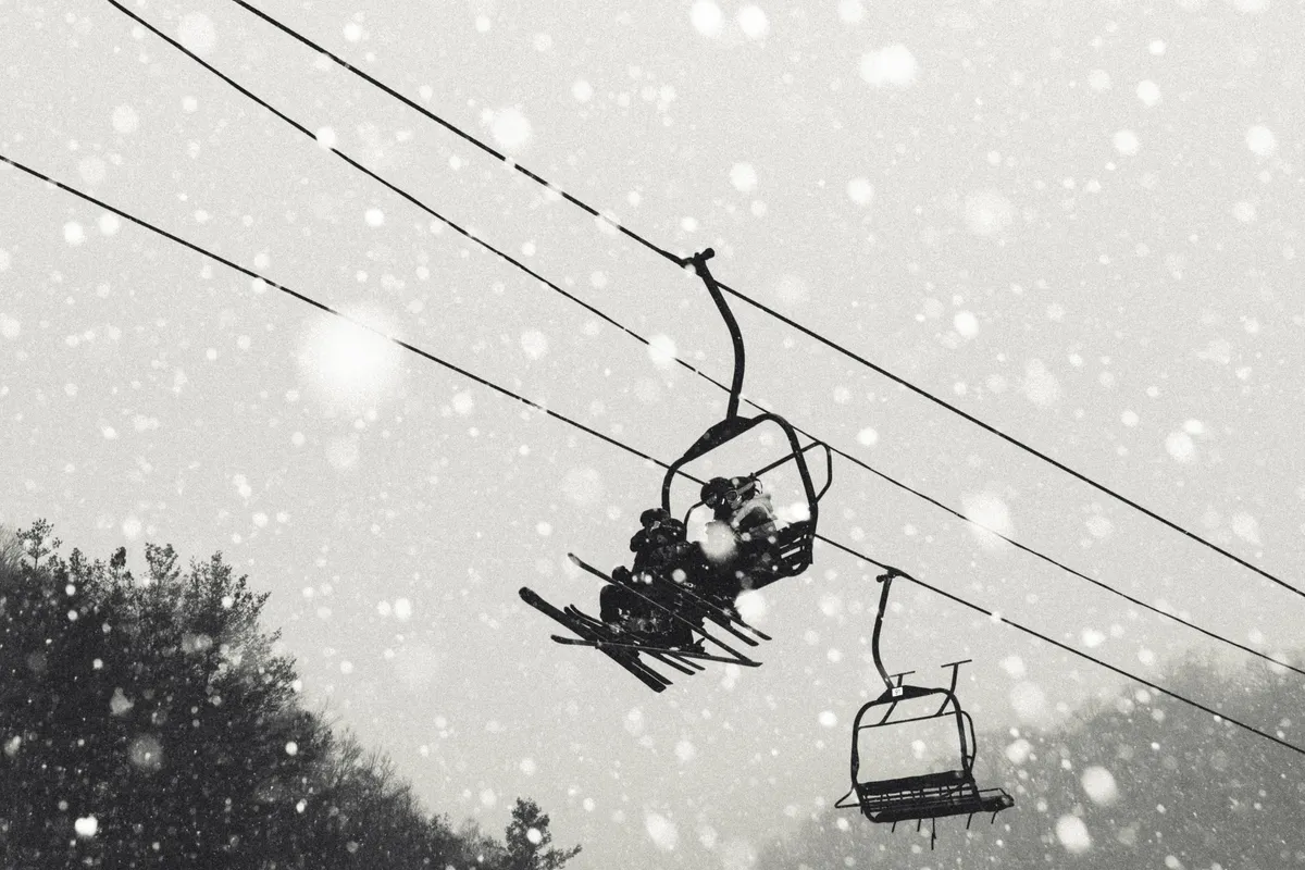 An exterior shot of a ski lift in motion, with snowflakes drifting down and trees in the background. The scene is serene yet evocative of the winter resort atmosphere, focusing on the infrastructure that makes skiing and snowboarding accessible.