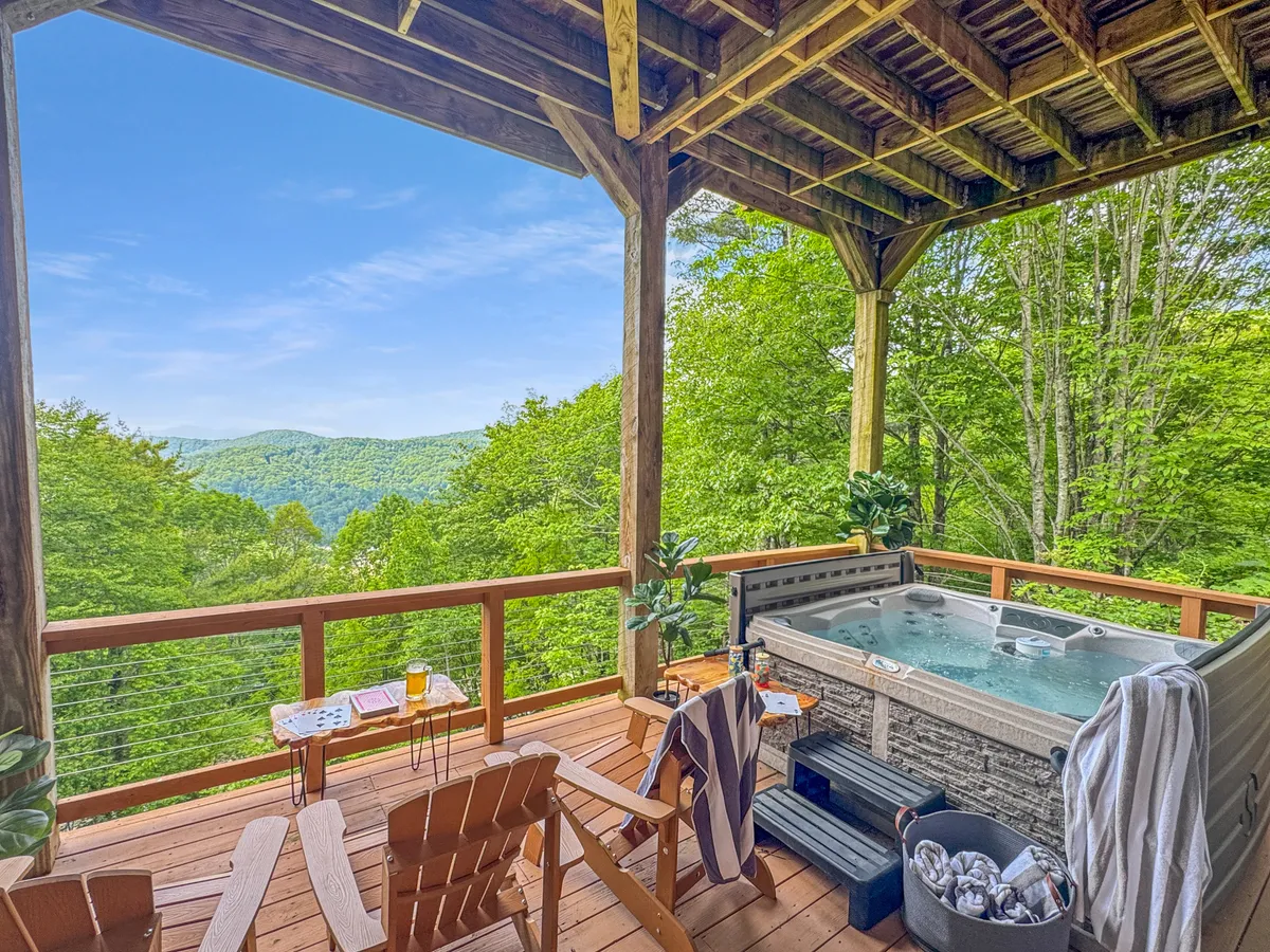 An inviting exterior shot of a hot tub set on a wooden deck overlooking a mountain view, with a comfortable seating area nearby. The space connects seamlessly to the outdoors and suggests relaxation after an active day.