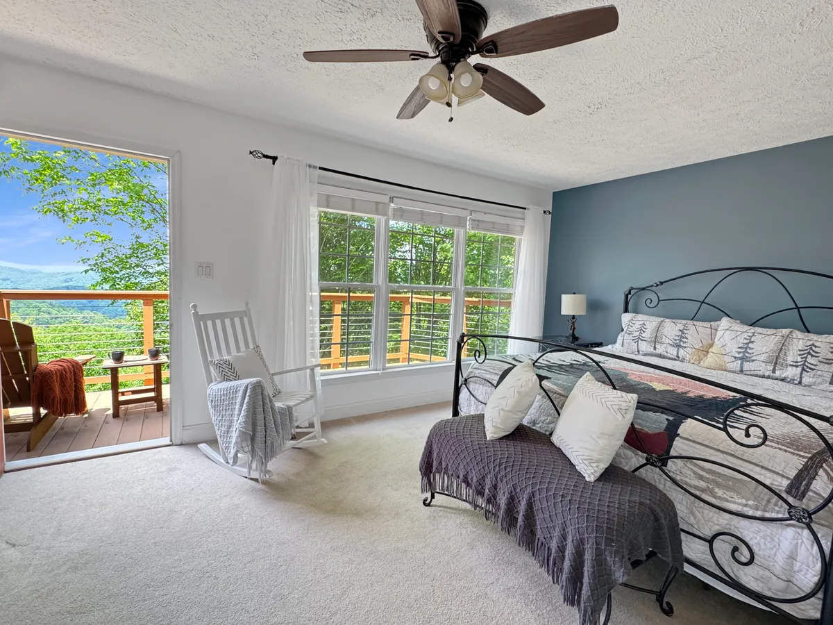 A serene bedroom with a plush bed adorned with decorative pillows, a large window letting in natural light, a cozy rocking chair, and a wooden deck visible outside. The ambiance is tranquil, with soft textures and a warm, inviting feel.