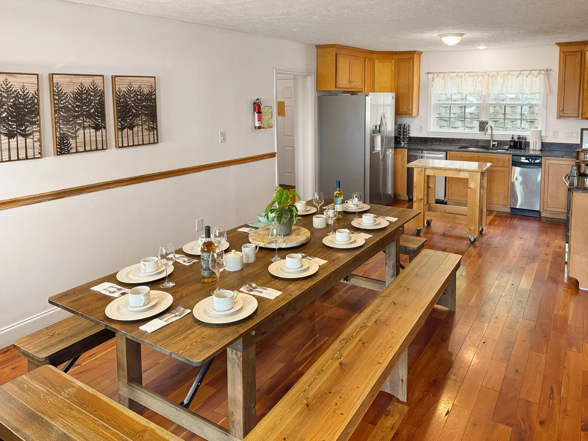 A large wooden dining table set for a meal, surrounded by comfortable chairs and warm decor elements like a decorative plant and framed artwork. Kitchen appliances are visible in the background, suggesting a fluid transition from dining to kitchen, ideal for group gatherings and shared meals.