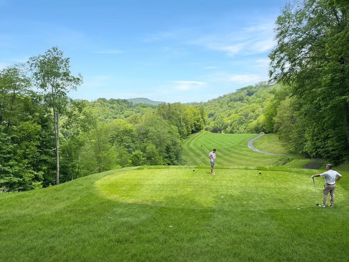 This image captures a group of golfers preparing to tee off on a lush, green course framed by rolling hills and mature landscaping. The wide, serene fairways and inviting natural setting reflect the leisurely and upscale country club experience.